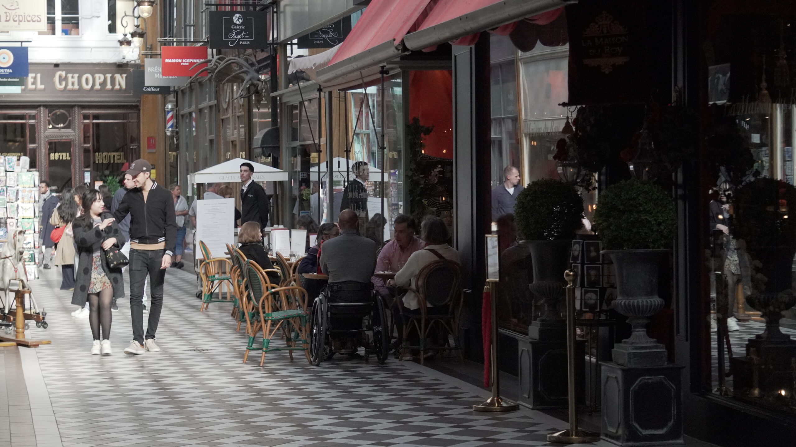 Terrasses de cafés animées dans le Passage Jouffroy à Paris, avec promeneurs et ambiance typique des passages couverts.